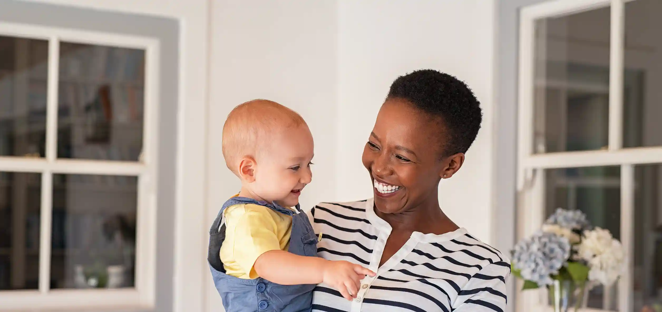 A happily smiling woman with dark skin tone, short dark hair and a striped shirt holds a happy infant with light skin.