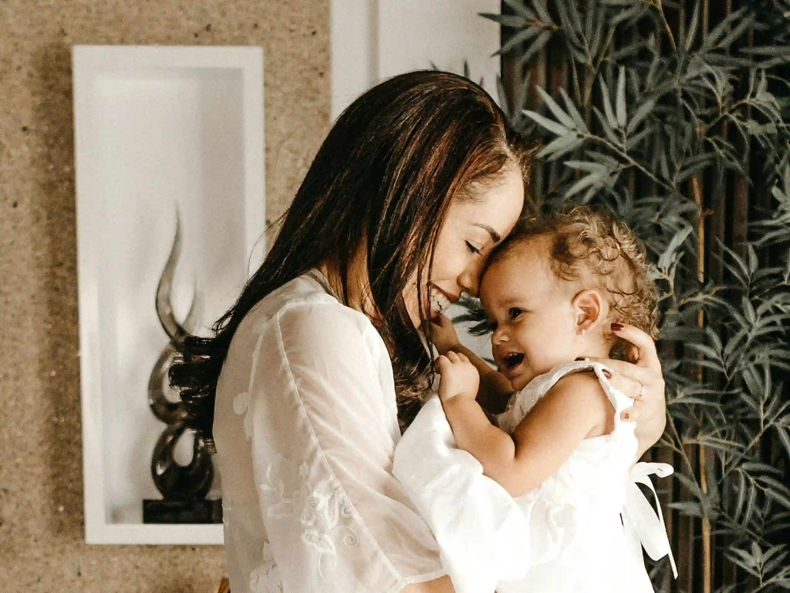 A mother and infant with light skin and medium hair smile happily while pressing their foreheads together.