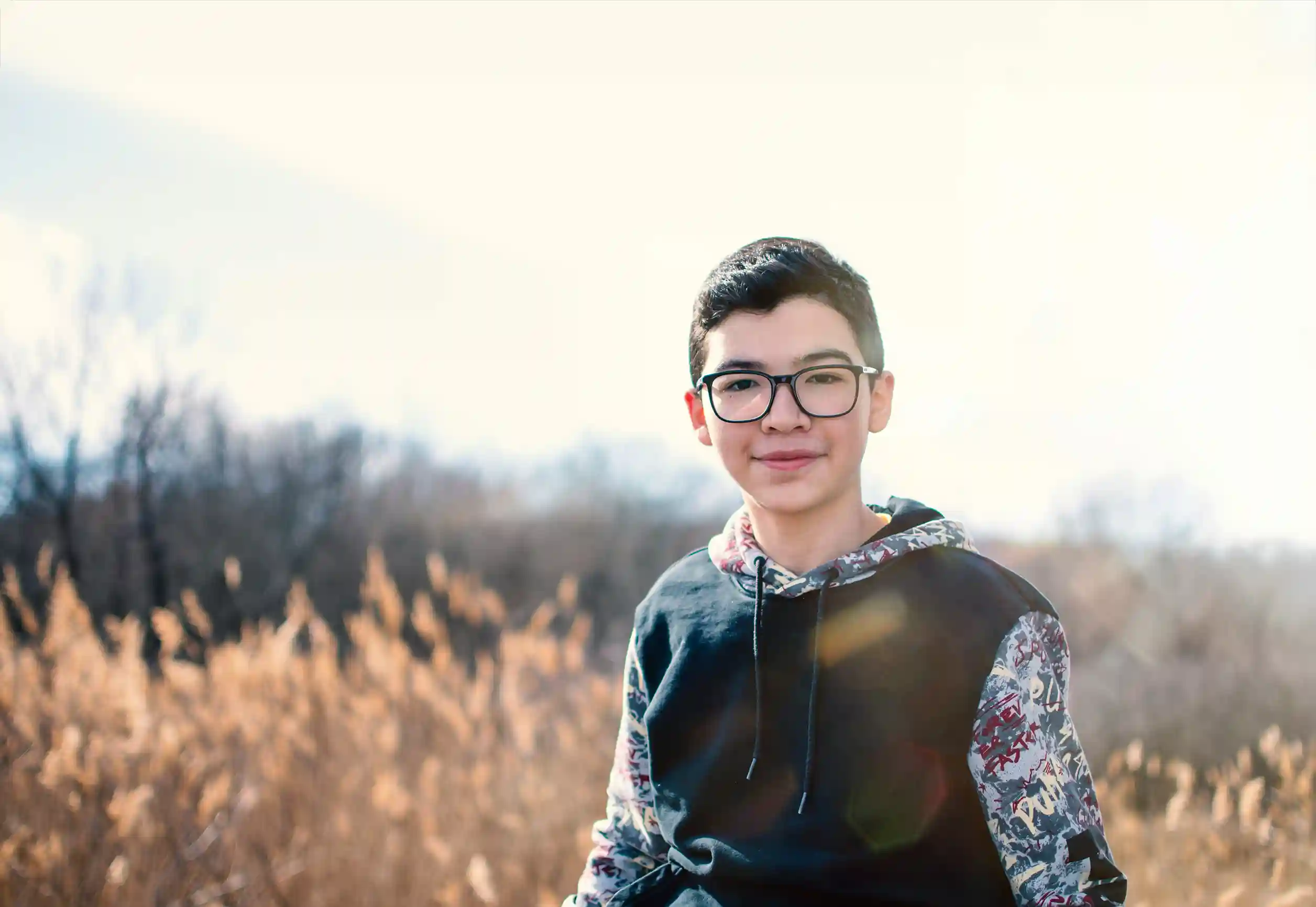 Teenager with short dark hair, wearing thick framed glasses and a black and patterned hoodie. They are in a field with tall grass, and are smiling and looking at the camera.