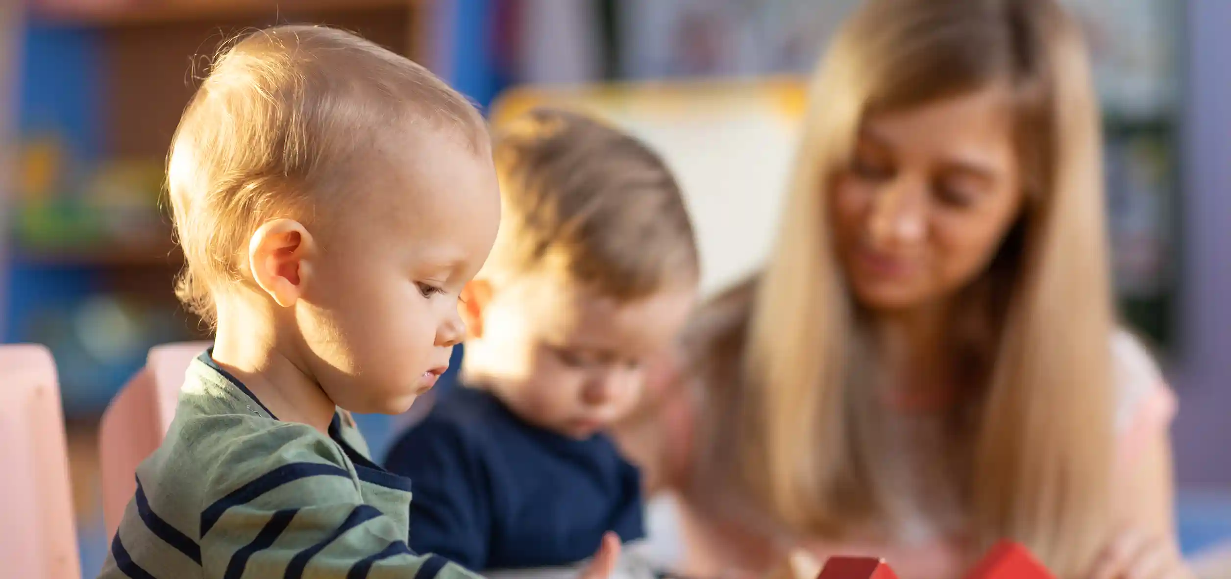 A light skinned toddler with blonde hair looks down at something, with another toddler and woman out of focus in the background.