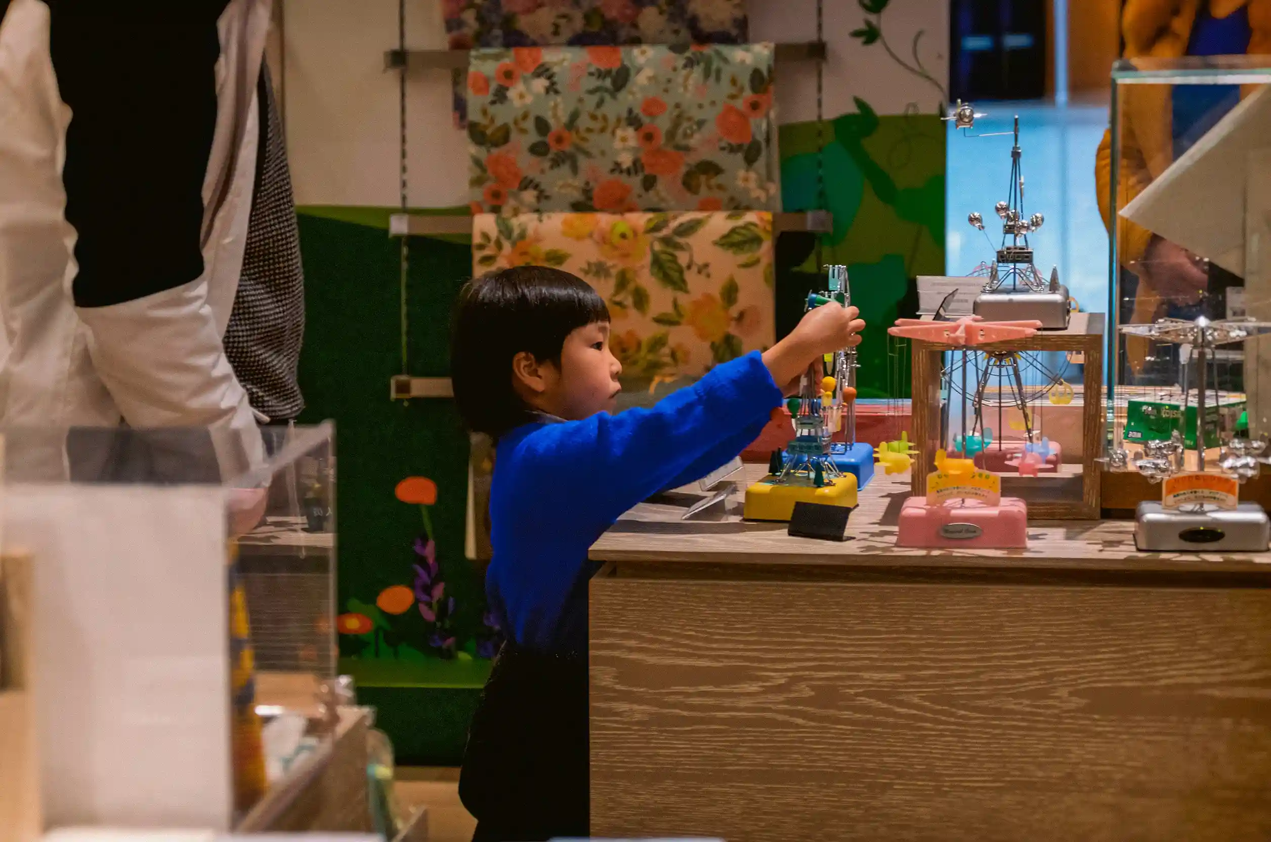 A toddler with medium skin tone, dark short hair and a dark blue sweater plays with toys on a table.