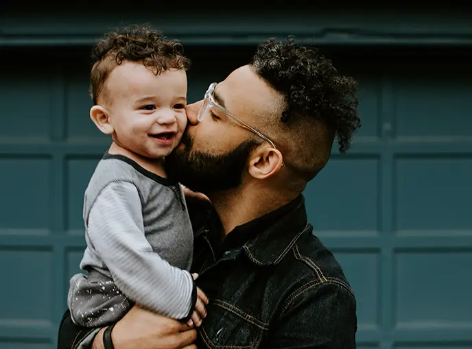 Hombre de mediana edad con tono de piel medio, pelo largo oscuro, gafas y una chaqueta de jean sostiene a su bebé sonriente en su pecho mientras le da un beso en la mejilla.