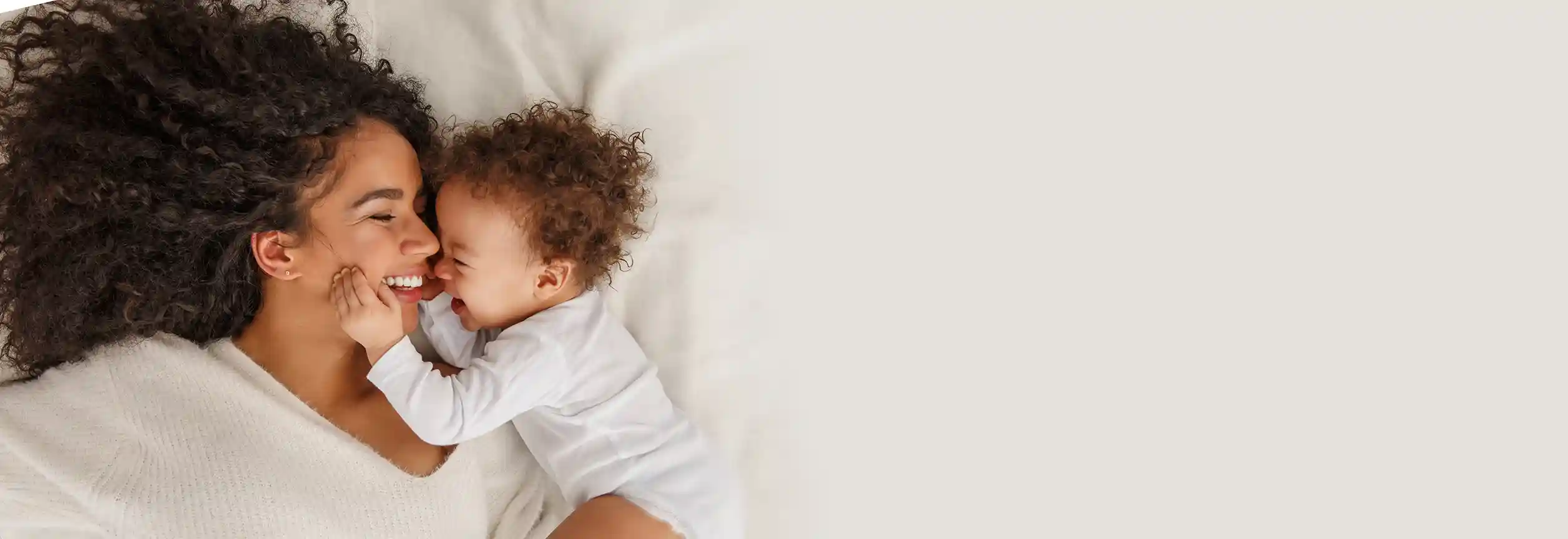 Una madre y un bebé de tono de piel medio-claro y pelo oscuro rizado yacen en la cama jugando; el bebé ríe y sostiene el rostro de la madre.