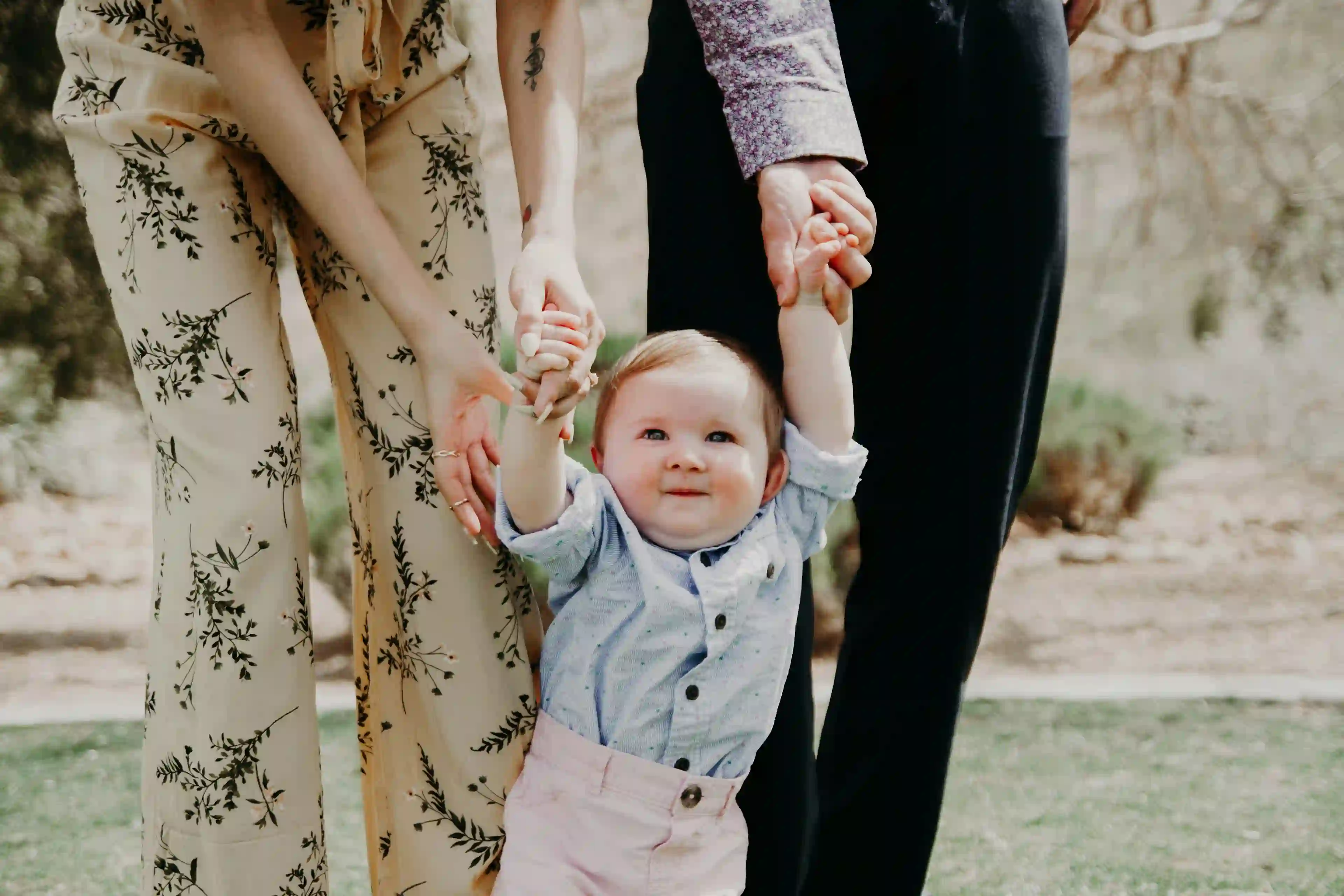 A light skinned infant with blonde hair walks while holding hands with parents that are out of frame.