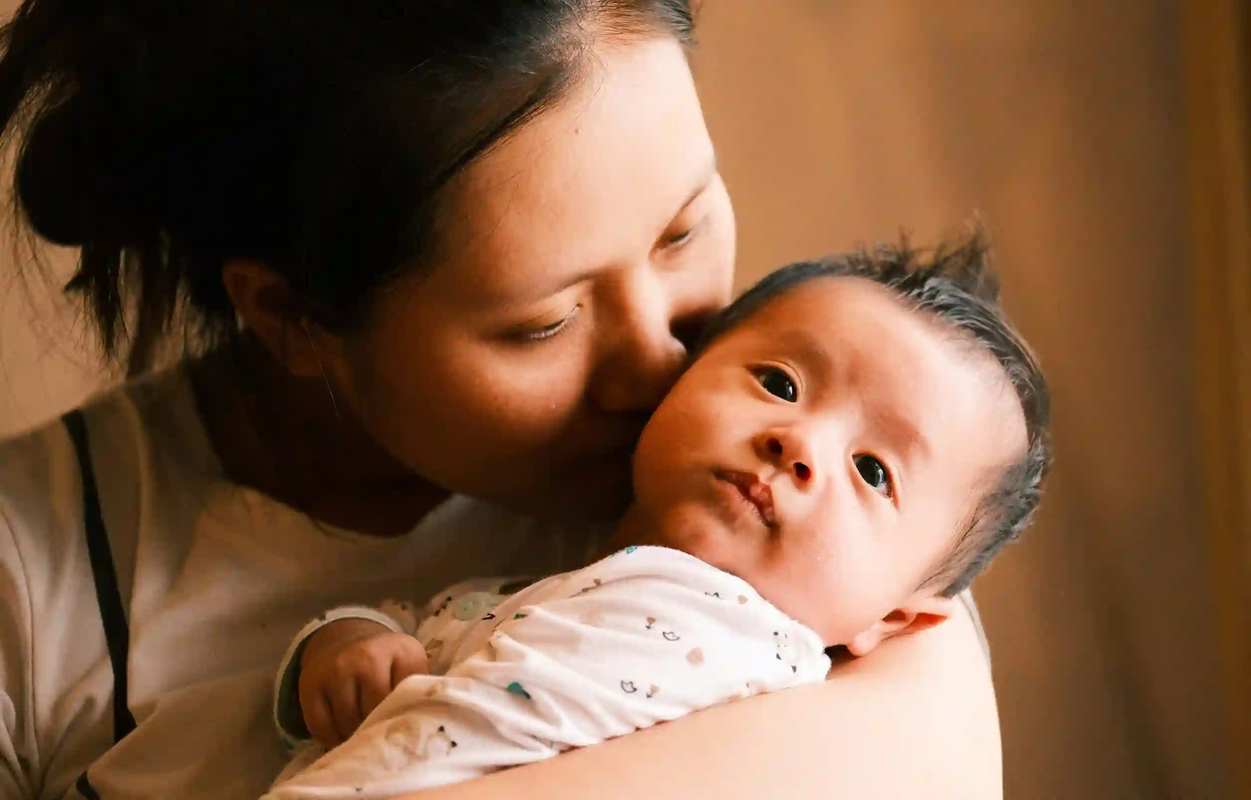 A medium-light skinned mother with long hair up in a bun kisses her infant on the cheek while holding them.