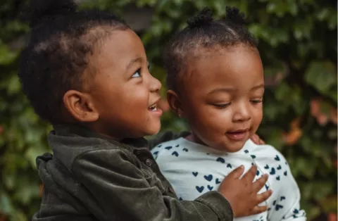 Two smiling children with dark skin tone embrace outside while looking away from the camera.