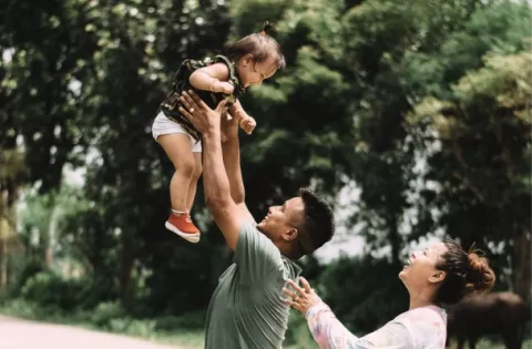 Una familia de piel de tono medio se ríe mientras el padre arroja a modo de juego a su hija al aire.