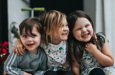 Three children with light skin tone sit on a step with arms around one another, smiling and giggling.