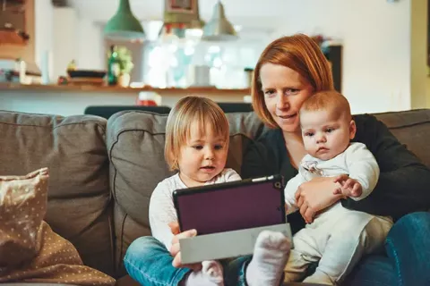 A woman with red hair and light skin tone holds her infant and toddler on her lap while they all look at a tablet.