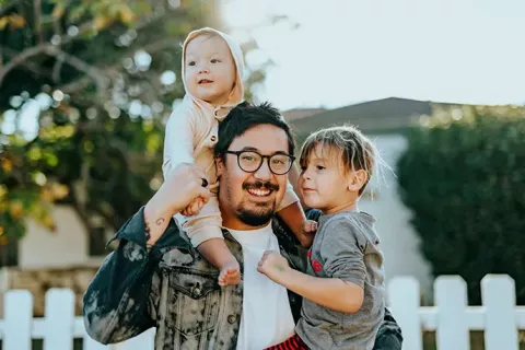 Un hombre sonriente con gafas, pelo corto y tono de piel medio-claro mira a la cámara mientras sostiene a un niño y un bebé con un tono de piel claro.