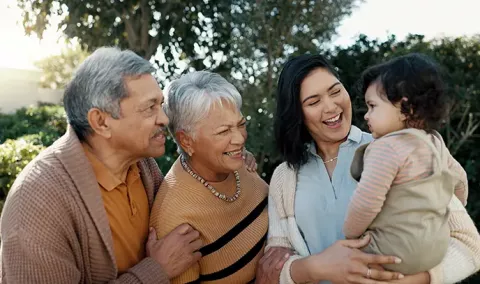 Una madre sonriente de cabello oscuro y tono de piel medio-claro sostiene a su niño pequeño mientras una abuela y un abuelo de edad avanzada se acercan a interactuar y jugar con el niño.